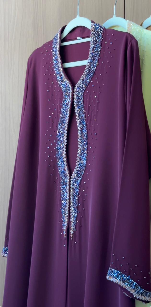 A long-sleeved purple abaya dress with sparkling silver and blue beaded trim along the V-neckline and cuffs, hanging on a white plastic hanger against a light wood-paneled wall.