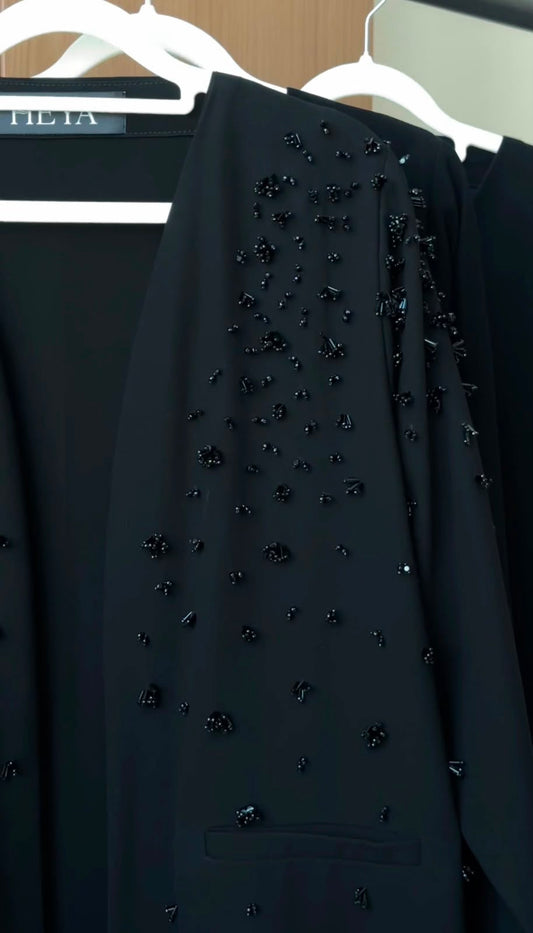 A close-up of a black tailored garment hanging on a white hanger, decorated with scattered dark beaded embellishments. Part of another black garment and a hanger with the partial brand name 'LILIA' are visible in the background.