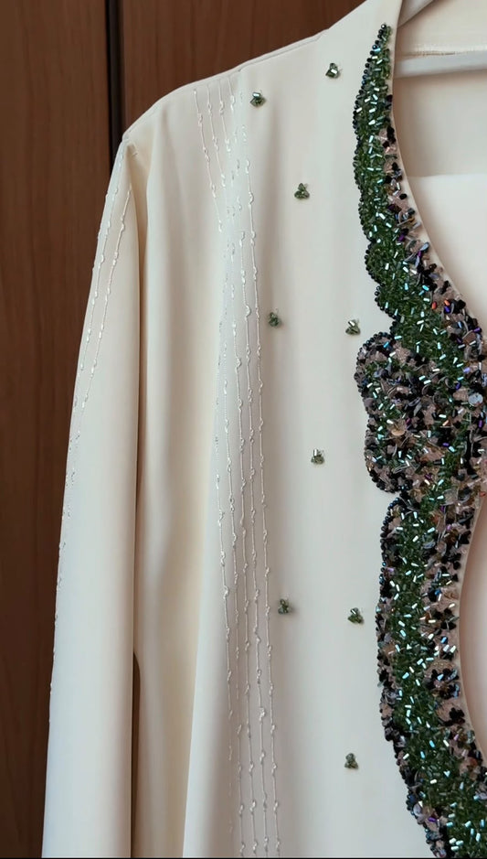 A cream-colored long-sleeve jacket or cardigan featuring delicate vertical bead embroidery on one side and a wide, scalloped border of green and silver sequins on the other, shown hanging against a wooden background.