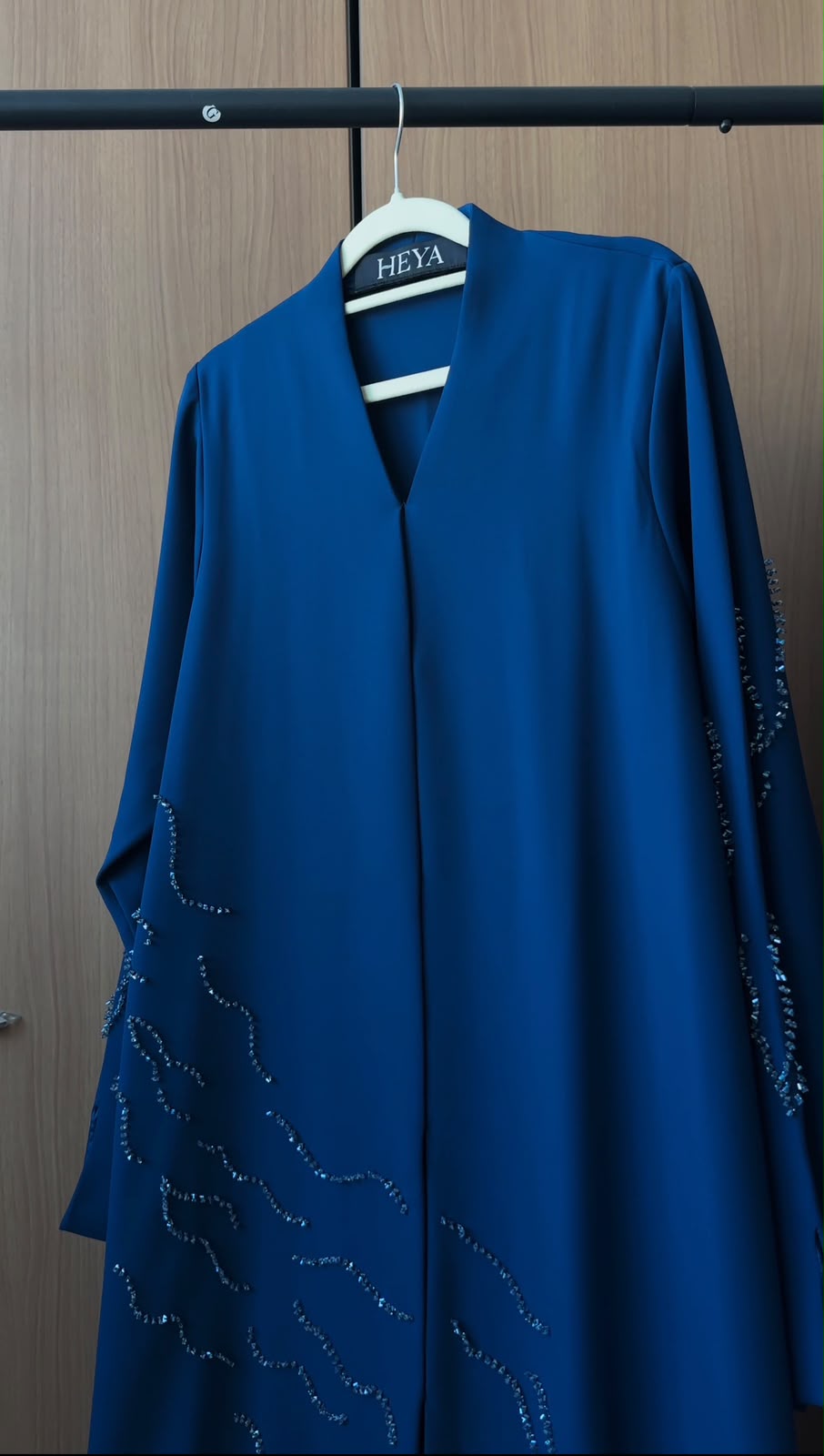 A royal blue abaya dress with subtle silver embroidered wave patterns on the lower front and sleeves, hanging on a white hanger labeled 'HEYA' against a brown wooden door background.