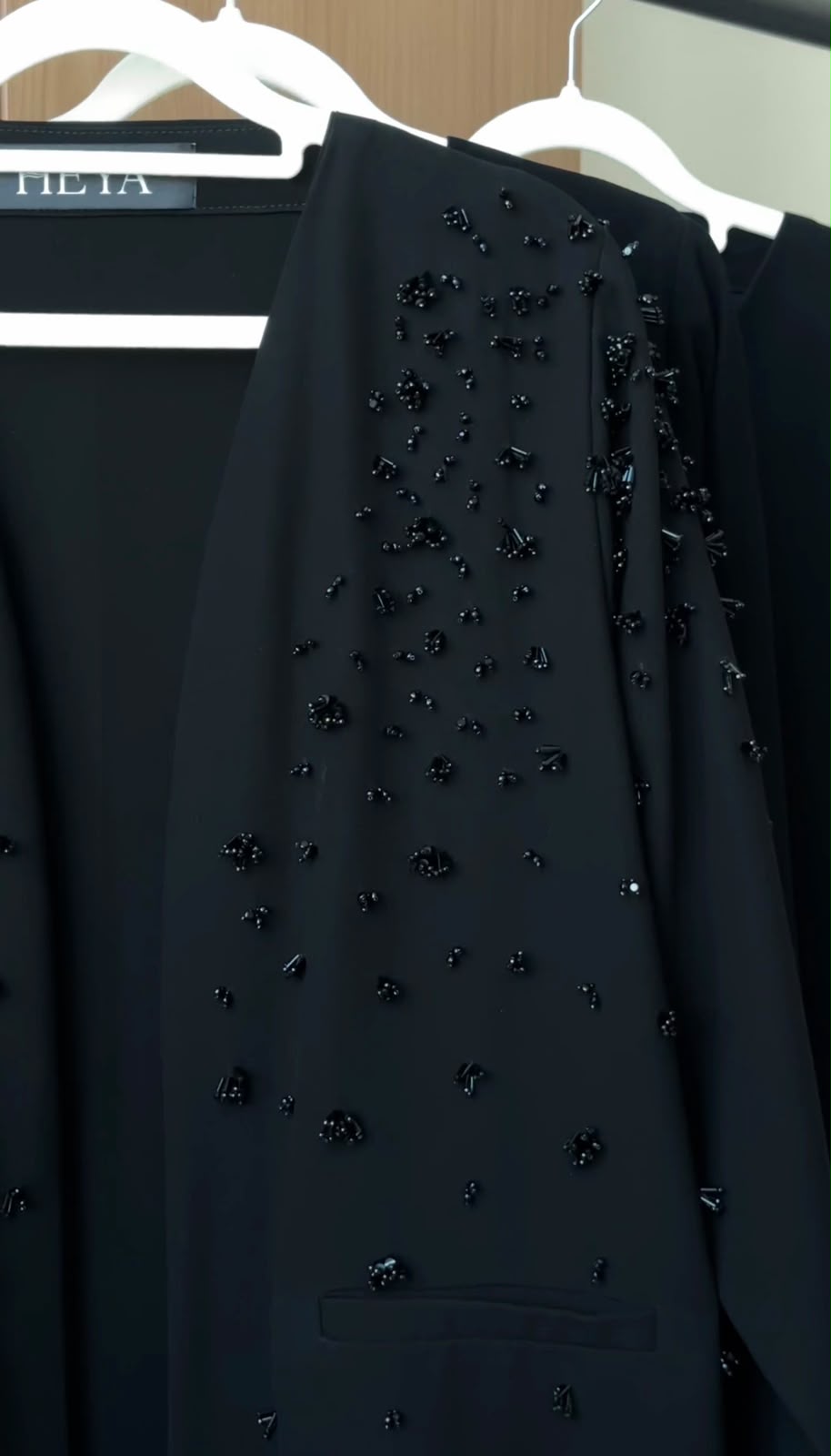 A close-up of a black tailored garment hanging on a white hanger, decorated with scattered dark beaded embellishments. Part of another black garment and a hanger with the partial brand name 'LILIA' are visible in the background.