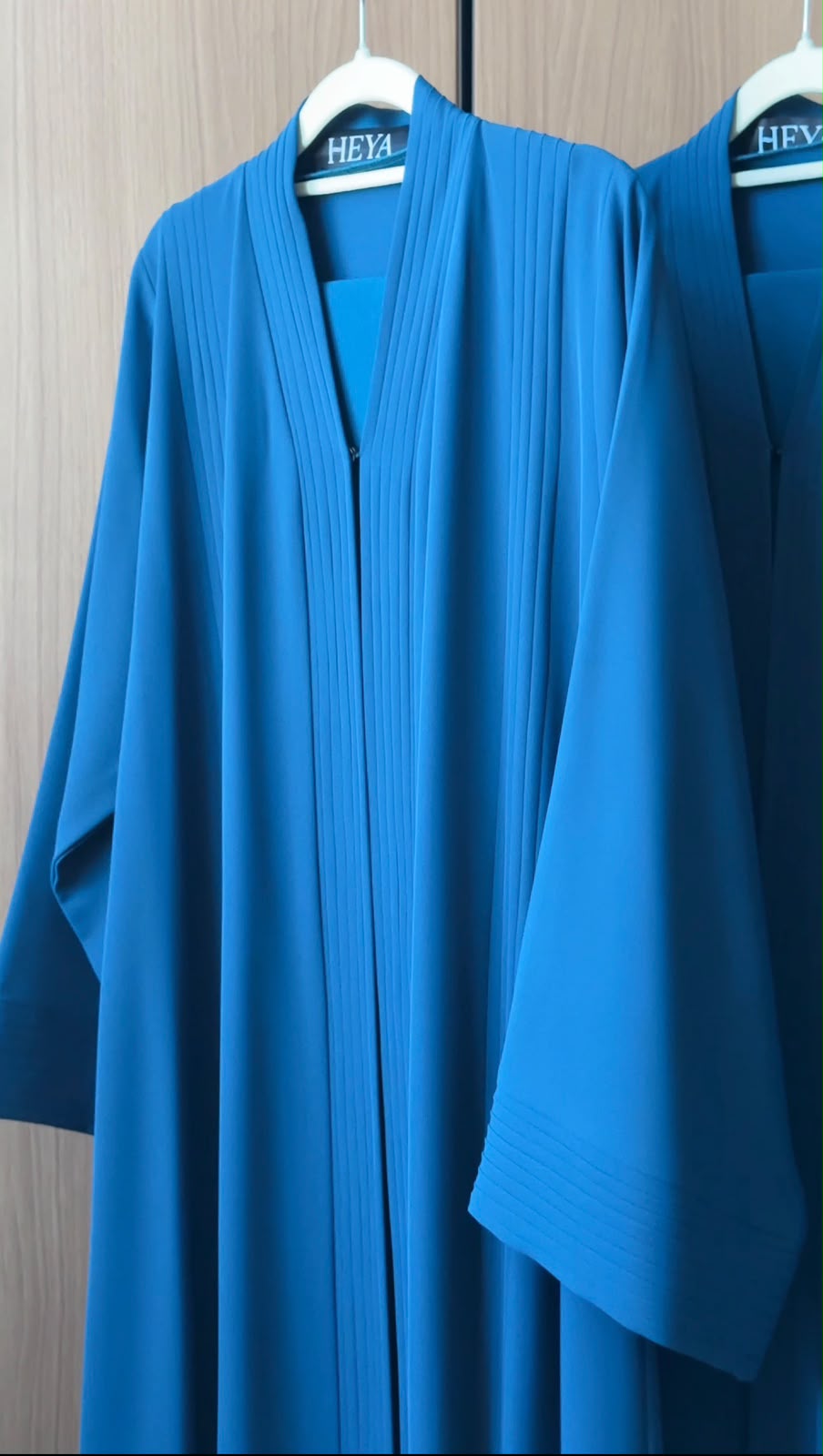 A bright blue kaftan robe with pleated detailing hangs on a white hanger against a light wood-paneled wall. The brand name 'HEYA' is visible on the neck label.