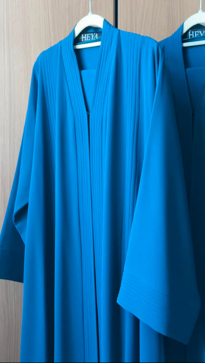 A bright blue kaftan robe with pleated detailing hangs on a white hanger against a light wood-paneled wall. The brand name 'HEYA' is visible on the neck label.