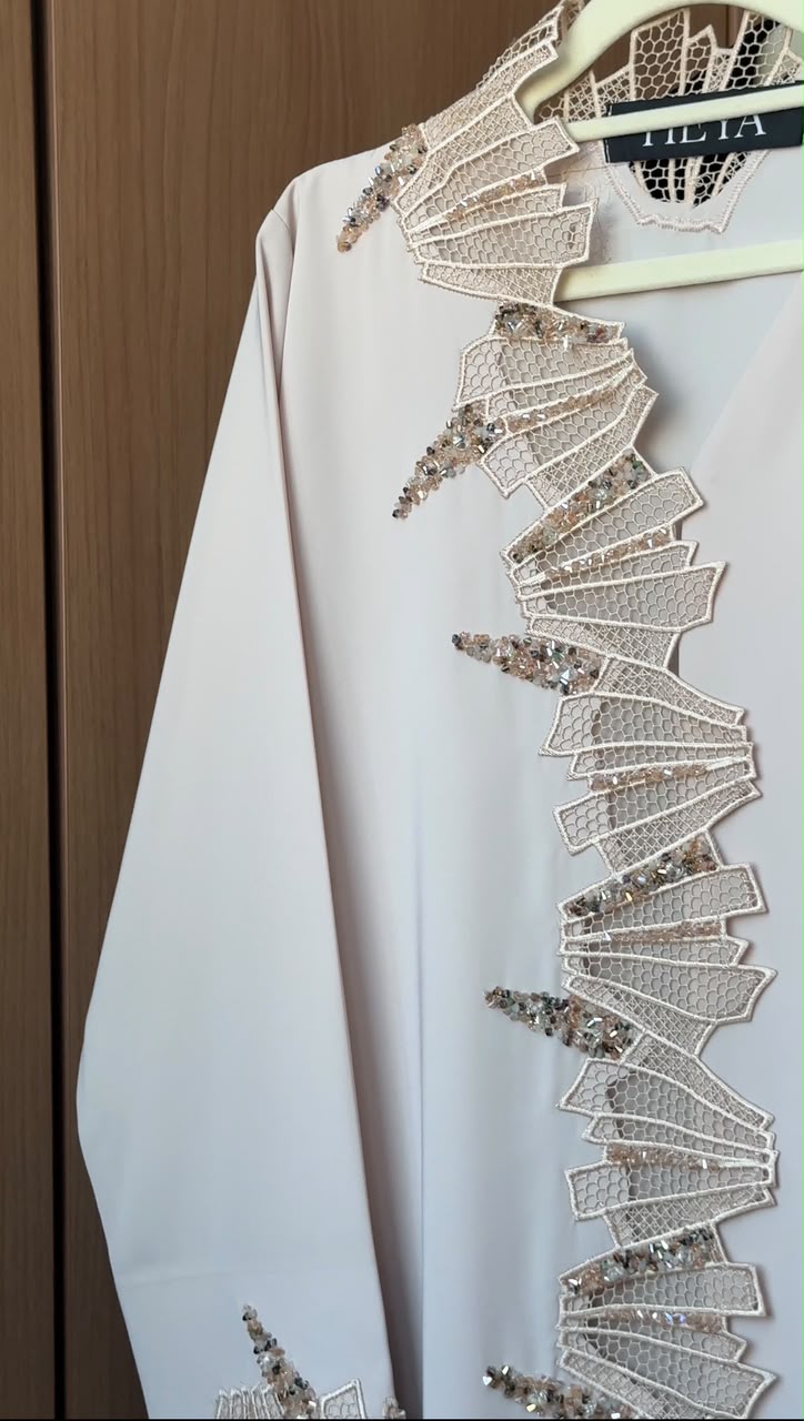 A white long-sleeve abaya with intricate beige and silver lace appliques and sequins along the front, hanging on a wooden hanger against a brown wooden door background.