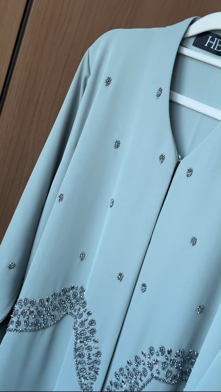 A light blue abaya jacket with silver rhinestone embellishments, hanging on a white hanger against a brown wooden background.