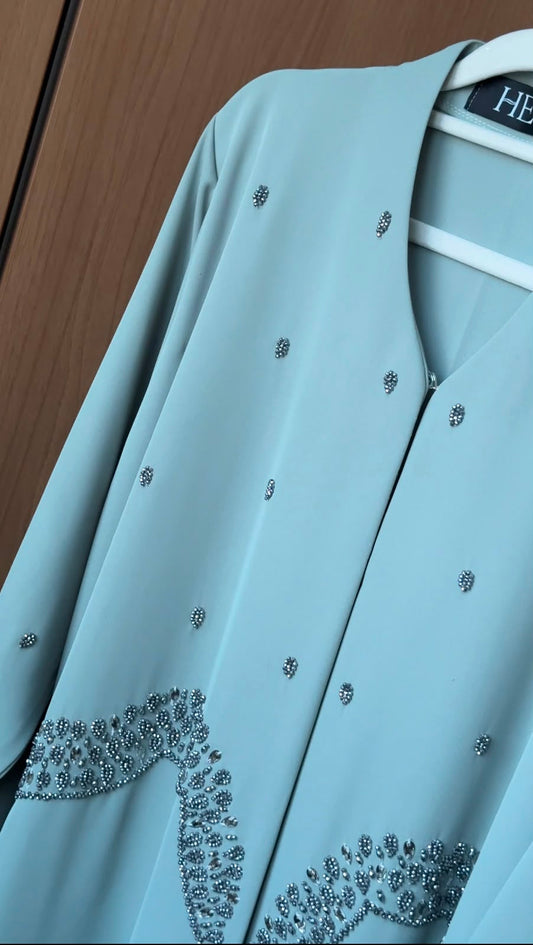 A light blue abaya jacket with silver rhinestone embellishments, hanging on a white hanger against a brown wooden background.