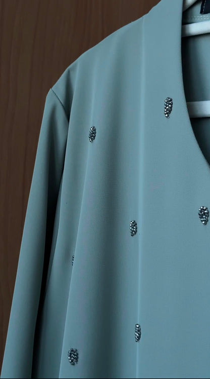 A sage green long-sleeve top with small silver rhinestone embellishments, hanging on a white hanger against a dark wood background.