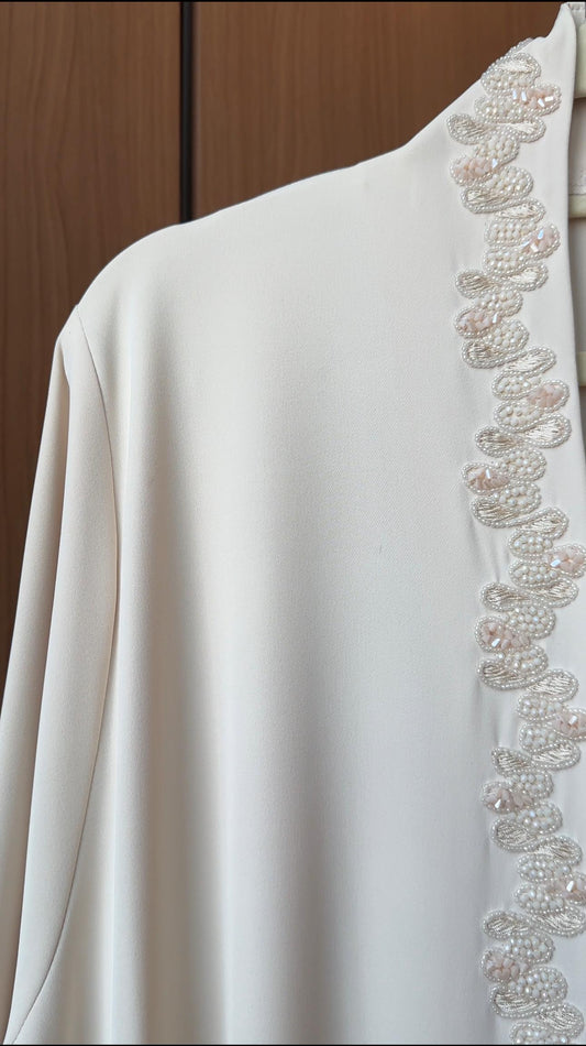 A close-up of a white or off-white garment with detailed beaded and sequined trim running down the shoulder and collar area, set against a dark wood background.