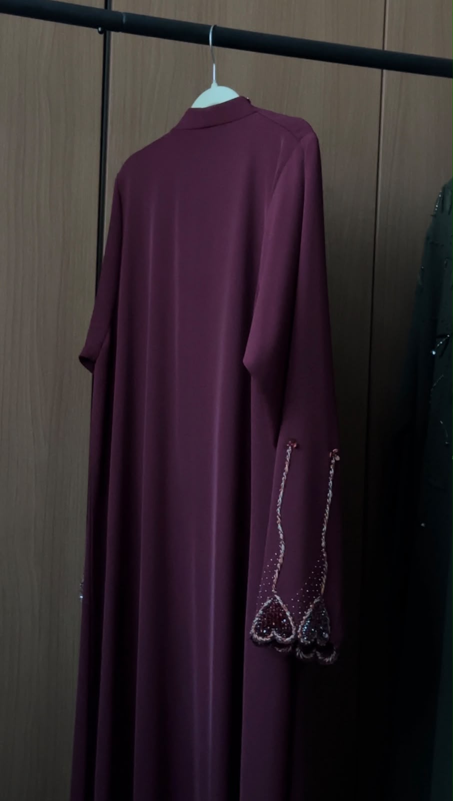 A long-sleeved, high-neck abaya dress in deep plum purple hanging on a white hanger against a dark wood-paneled wall. The sleeves feature decorative silver beaded fringe detailing near the cuffs.