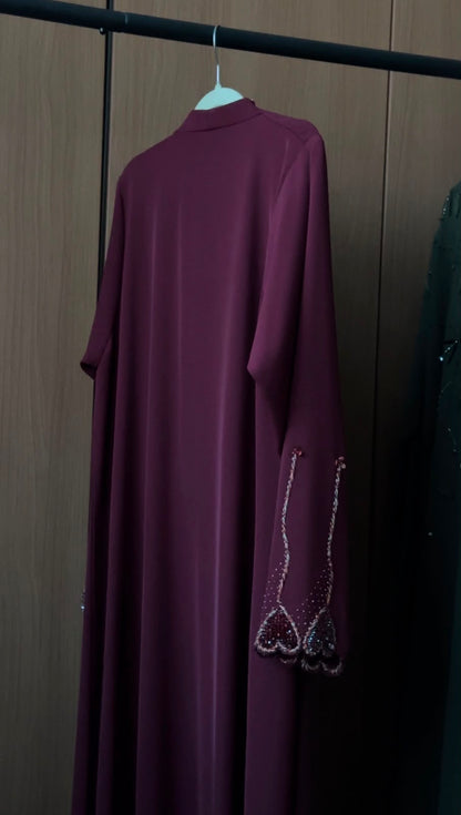 A long-sleeved, high-neck abaya dress in deep plum purple hanging on a white hanger against a dark wood-paneled wall. The sleeves feature decorative silver beaded fringe detailing near the cuffs.
