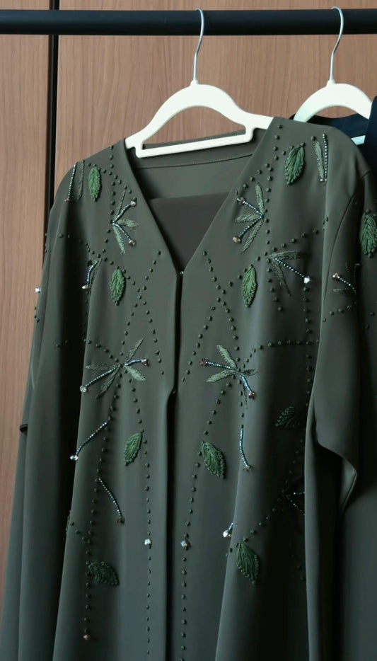 A dark olive green long-sleeve abaya with intricate embroidered dragonflies and leaf patterns, hanging on a white hanger against a dark wood background.