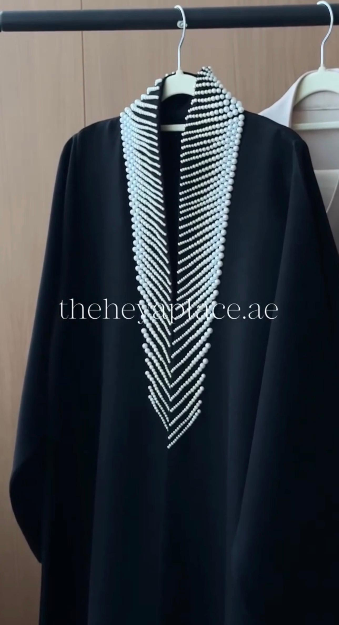 A black abaya or long robe hanging on a hanger, featuring a wide V-shaped neckline embellished with multiple strands of white pearls. The garment is shown against a neutral indoor background with a wooden door and another hanger partially visible.
