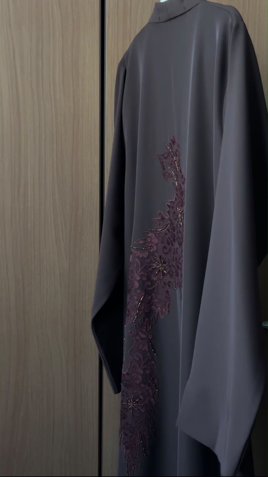 A dark gray abaya with a vertical plum-colored embroidered pattern on the side, hanging against a wooden door background.