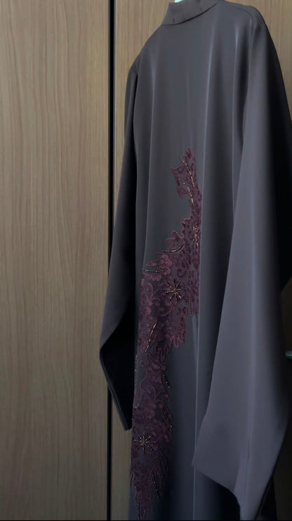 A dark gray abaya with a vertical plum-colored embroidered pattern on the side, hanging against a wooden door background.