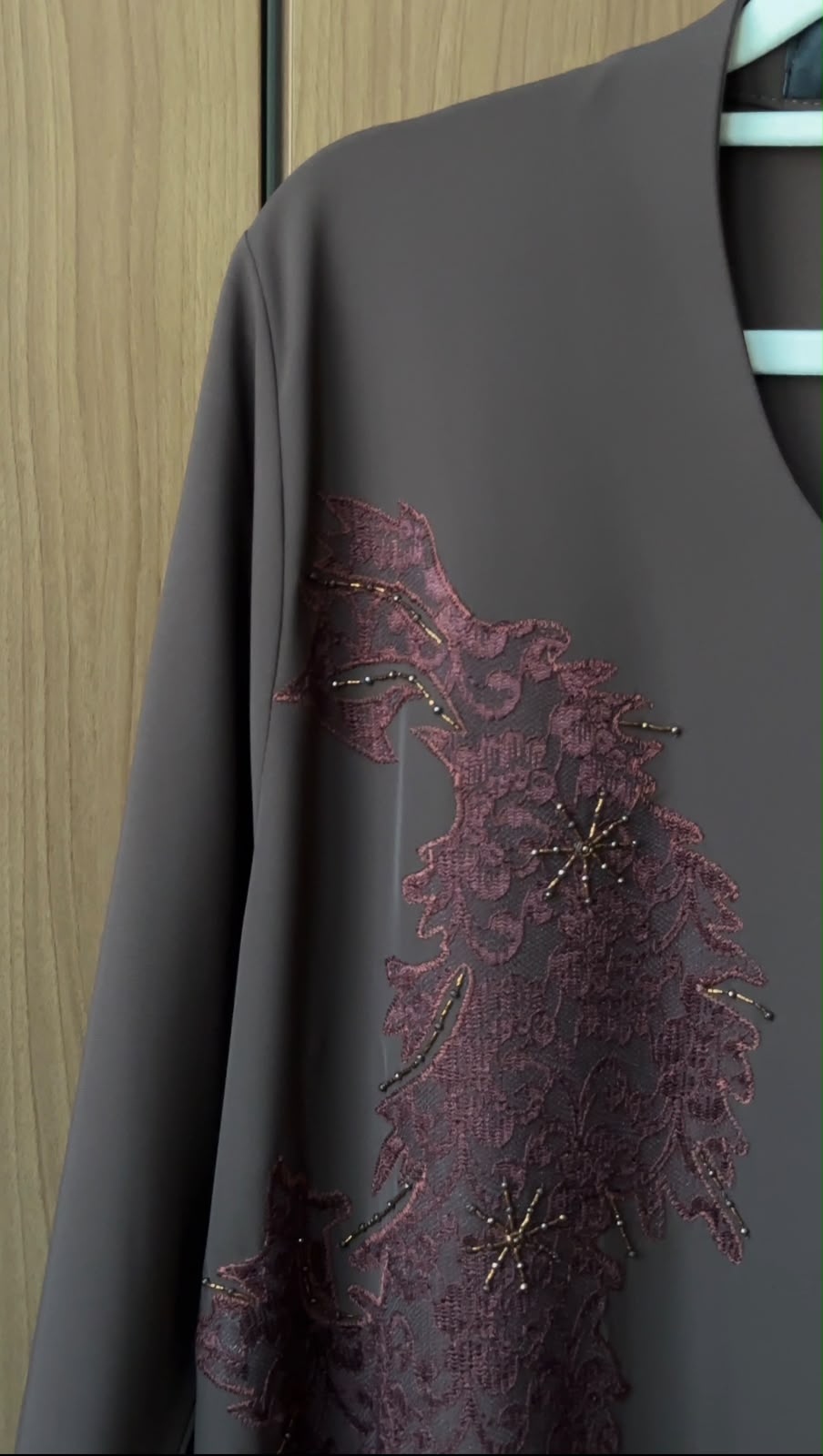 A dark gray long-sleeve top or dress featuring a large burgundy lace applique panel with gold beaded star accents on the front, hanging on a light blue hanger against a wooden door background.