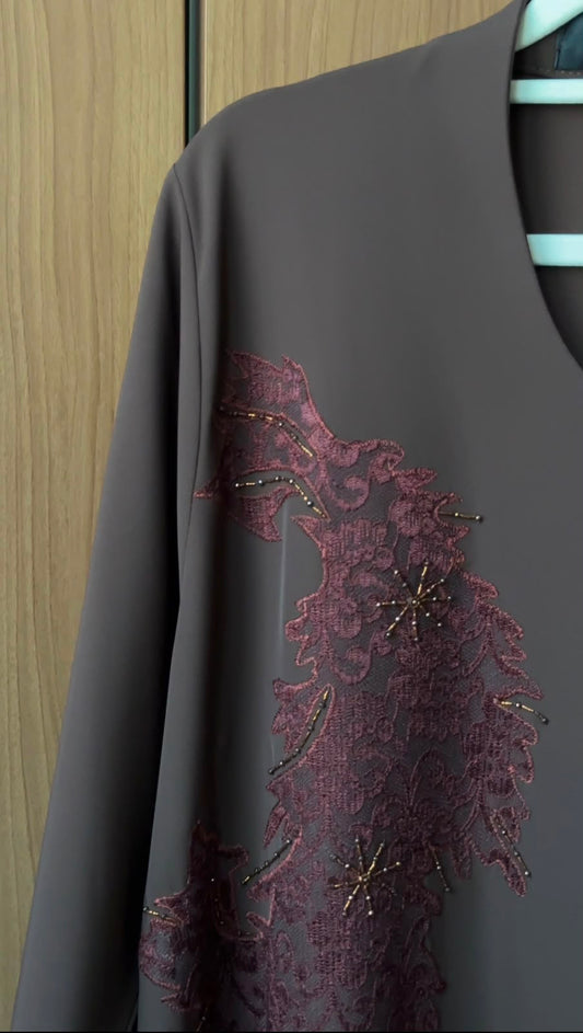 A dark gray long-sleeve top or dress featuring a large burgundy lace applique panel with gold beaded star accents on the front, hanging on a light blue hanger against a wooden door background.