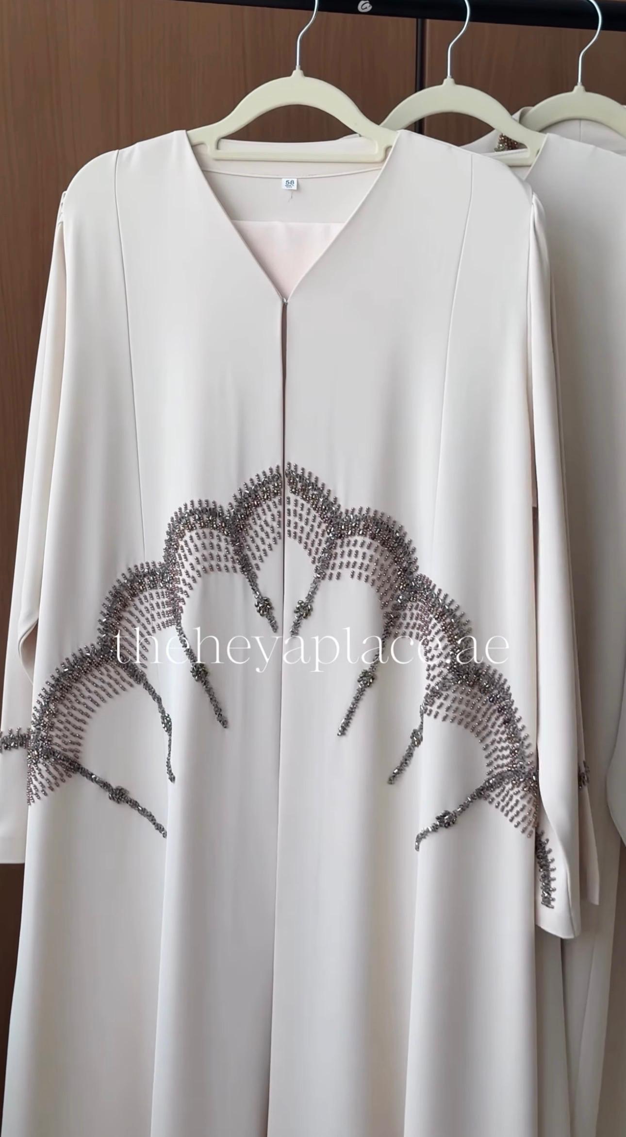 A cream-colored long abaya dress with a V-neckline and a scalloped, embroidered fan-like pattern across the lower front, hanging on a wooden hanger.