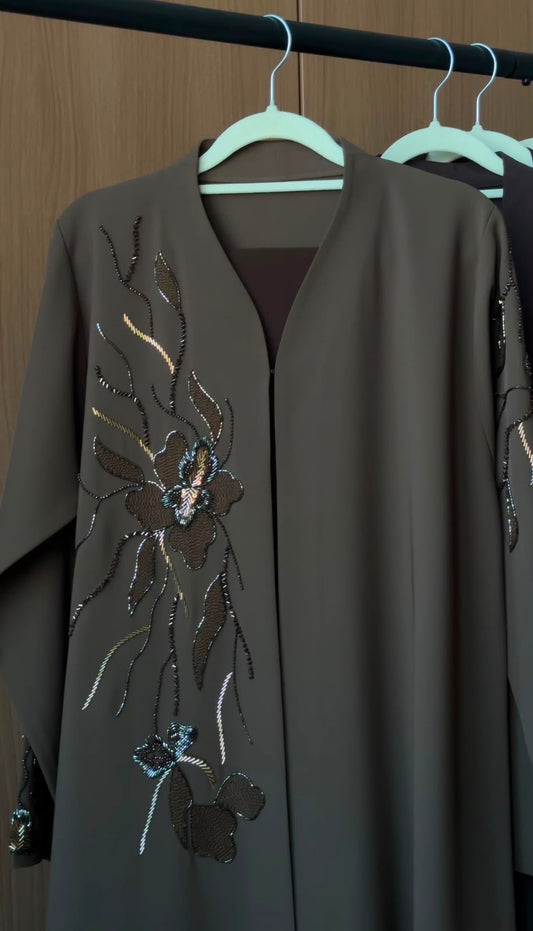 A dark brown abaya with intricate floral sequin embroidery on the front, hanging on a white hanger on a clothing rack against a wooden background.
